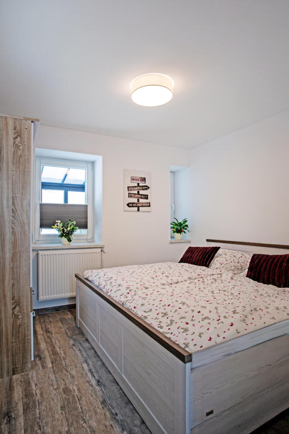 Ein helles Schlafzimmer mit einem großen Bett, Bettwäsche mit Blumenmuster, zwei Fenstern, Pflanzen auf den Fensterbänken, einem Kleiderschrank aus Holz und einer Deckenlampe. Die Wände sind weiß und der Boden hat eine Holzoptik.
