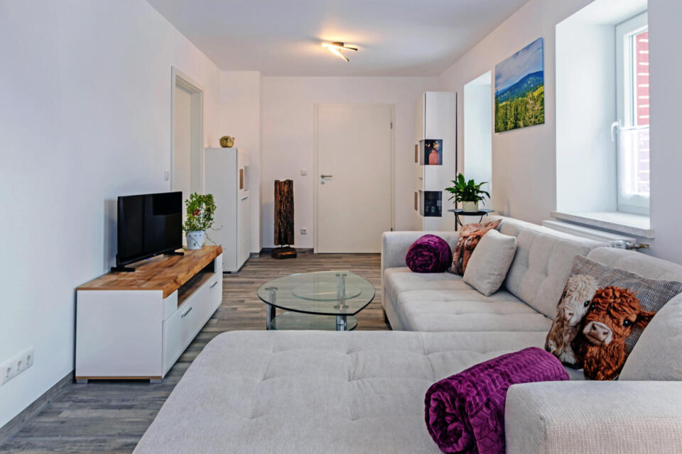 Modernes Wohnzimmer mit einem großen hellgrauen Ecksofa, violetten Kissen, einem Couchtisch aus Glas, einem Fernseher auf einem Holzständer, Zimmerpflanzen und Wandbildern neben zwei Fenstern. Der Boden besteht aus Holz, die Wände sind weiß.