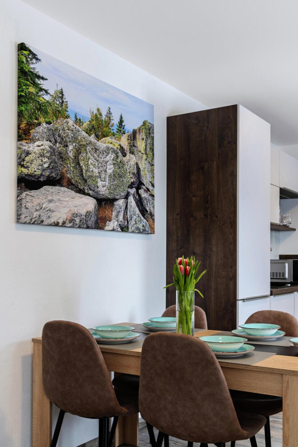 Ein moderner Essbereich mit vier braunen Stühlen, die um einen Holztisch herum stehen, auf dem grüne Teller und eine Vase mit roten Tulpen stehen. An der weißen Wand über dem Tisch hängt ein großes Naturfoto mit Felsen und Bäumen.