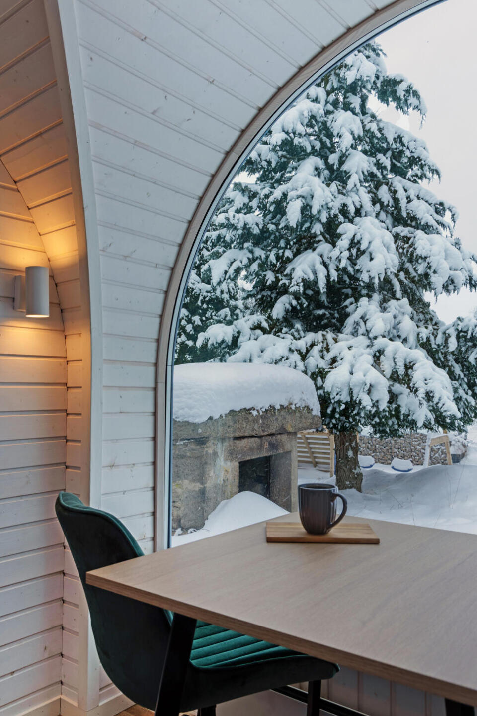 Durch ein geschwungenes Fenster bietet sich der Blick auf eine verschneite Landschaft mit einem großen, schneebedeckten Baum. Im Innenraum steht ein Holztisch mit einem Becher und einem Stuhl neben weiß getäfelten Wänden, die von einer Wandlampe in warmes Licht getaucht werden.