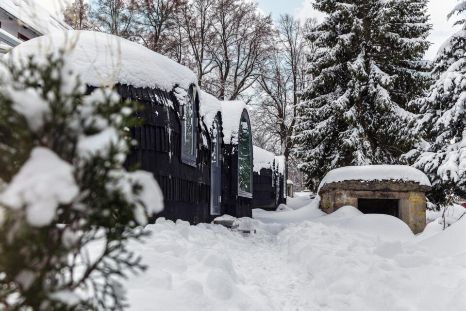 Geschwungene schwarze Hütten und ein Steingebäude liegen inmitten von tiefem Schnee und hohen Bäumen, die alle dick mit Neuschnee bedeckt sind, und bilden so eine friedliche Winterlandschaft.