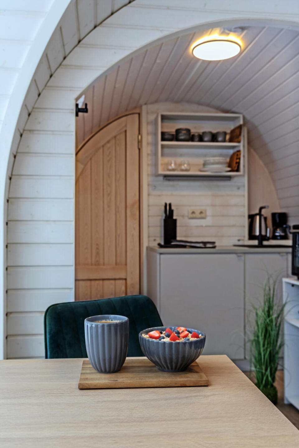 Auf einem Holztablett, das auf einem Tisch steht, stehen eine graue Tasse und eine Schale mit bunten Bonbons, im Hintergrund ist eine gemütliche, moderne Küche mit einem gewölbten Holztürrahmen zu sehen.