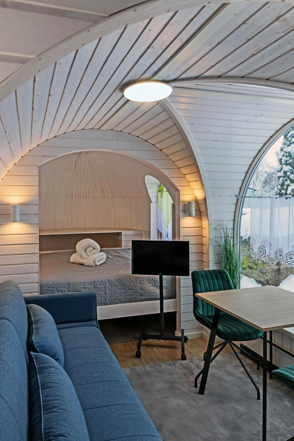 Modernes Tiny-House-Interieur mit geschwungenen Holzwänden, einem gemütlichen Bett, einem blauen Sofa, einem kleinen Fernseher auf einem Ständer, einem grünen Sessel und einem runden Fenster, durch das man eine verschneite Landschaft im Freien sehen kann. Neutrale Farbtöne und sanfte Beleuchtung sorgen für eine warme Atmosphäre.
