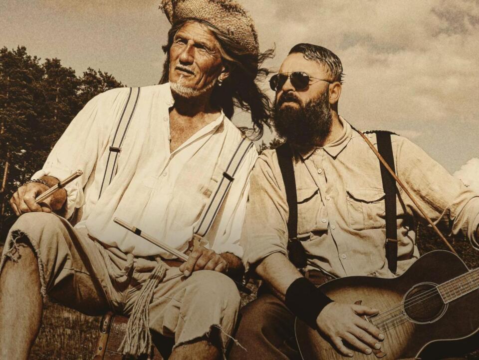 Zwei Männer sitzen im Freien und tragen robuste Kleidung im Vintage-Stil in einer „After Work Siebenquell“-Kulisse. Der eine hält Trommelstöcke in der Hand und trägt einen Hut mit Hosenträgern, während der andere einen Bart und eine Sonnenbrille trägt und Gitarre spielt. Das Bild ist in Sepia-Tönen gehalten, was ihm einen altmodischen Look verleiht.