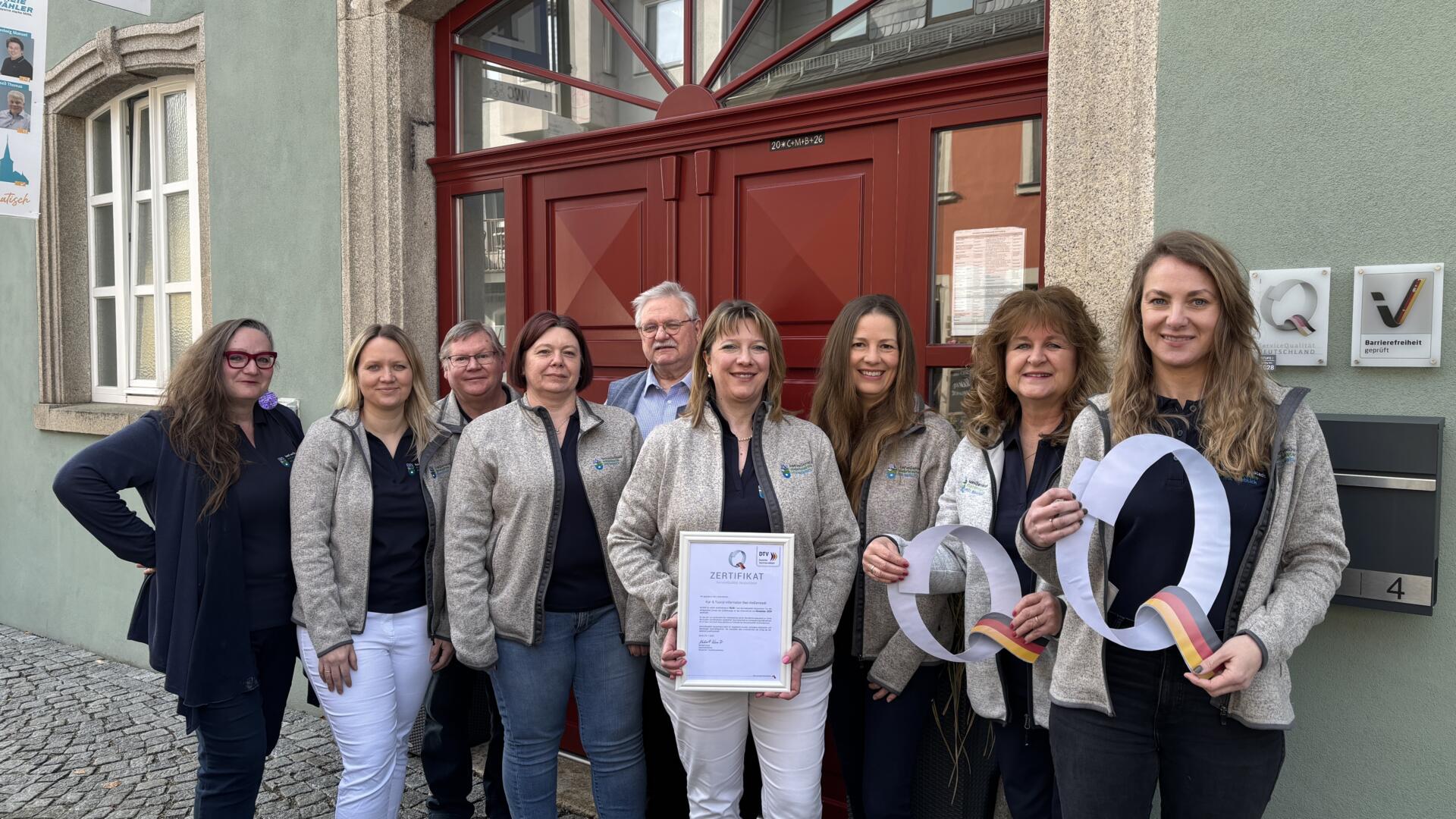 Acht Personen stehen vor dem Gebäude der Kur- und Touristeninformation, einige von ihnen lächeln in die Kamera. Eine Person hält eine Urkunde in der Hand, während zwei andere große, weiße, ausgeschnittene „Q“-Formen hochhalten. Im Hintergrund sind eine rote Tür und Schilder zu sehen.