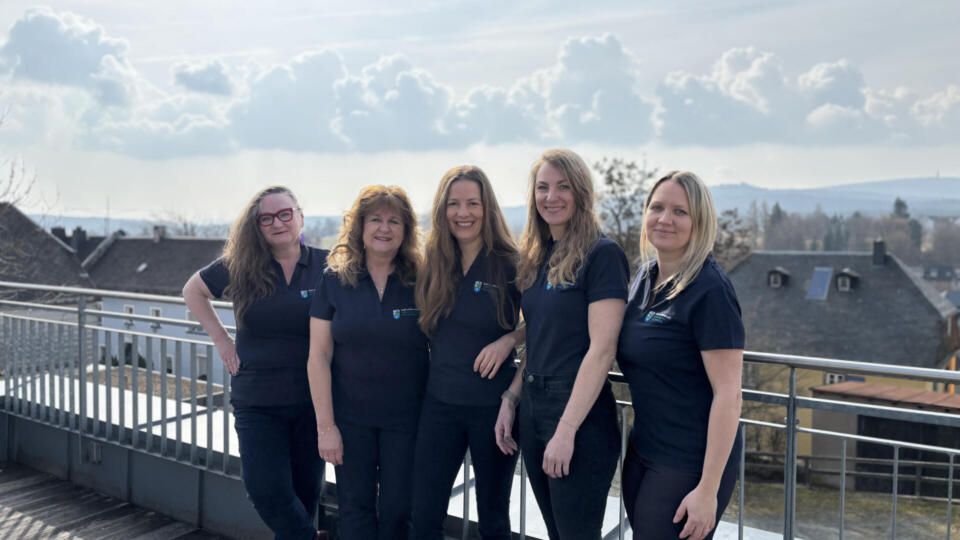 Fünf Frauen in einheitlichen marineblauen Shirts stehen nebeneinander und lächeln auf einer Außenterrasse mit Geländer. Als Vertreterinnen des Teams der Kur- und Tourist-Information posieren sie vor bewölktem Himmel, im Hintergrund sind in der Ferne Gebäude zu sehen.